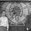 [Ann Steinweg and an Andrews University faculty member stand beside a mural of her own making. The mural depicts Christ's hand holding the symbol of Andrews University]