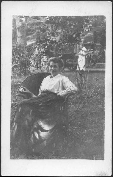 M. Bessie DeGraw sitting in wicker chair eating an apple, smiling at camera