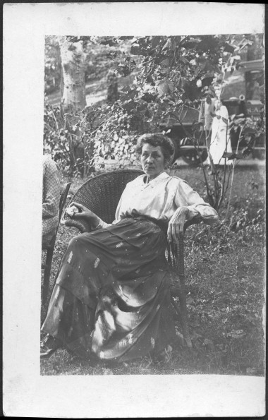 M. Bessie DeGraw sitting in wicker chair eating an apple