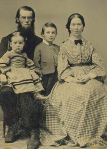 Mostly monochromatic image of a bearded man seated, woman in a long dress seated, young boy standing, and a young girl sitting on the man's lap.