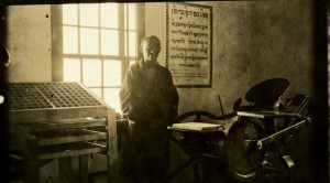 Mans standing beside window, and next to a printing press, and boxes that look like they hold lead type.