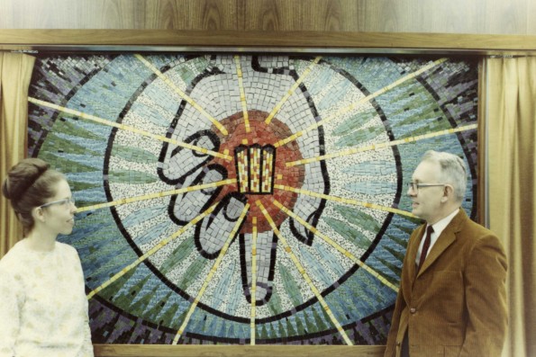 [Ann Steinweg and an Andrews University faculty member stand beside a mural of her own making. The mural depicts Christ's hand holding the symbol of Andrews University]